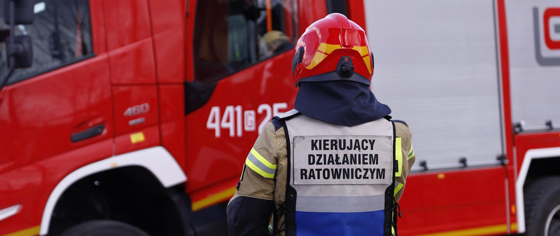 Na zdjęciu widać strażaka, kierującego działaniem ratowniczym. W tle widoczny samochód pożarniczy.