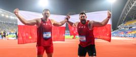 Na środku wielkiego stadionu dwóch mężczyzn w czerwonych sportowych ubraniach trzyma polskie flagi.