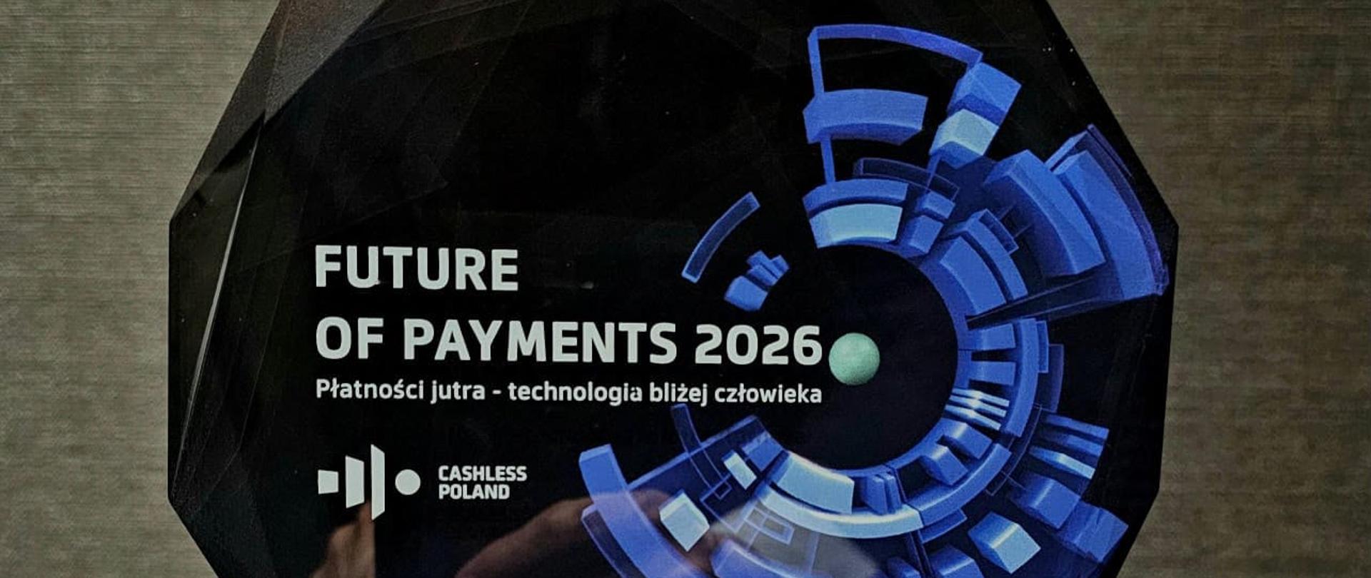 Statuetka „Future of Payments 2026” ustawiona na drewnianej podstawie. Na froncie widoczny jest napis „Future of Payments 2026”, logo Cashless Poland oraz niebieska grafika technologiczna. Na tabliczce podstawy znajduje się napis „Wydarzenie przełomowe na rynku, Warszawa 2026”.