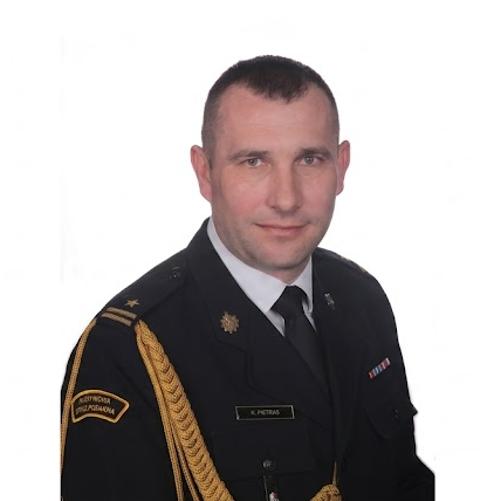 Portret mężczyzny w ciemnym mundurze Państwowej Straży Pożarnej, przedstawiony w bardzo szerokim formacie panoramicznym na białym tle. Mężczyzna ma krótkie, ciemne włosy i patrzy prosto w obiektyw z lekkim uśmiechem. Na lewym ramieniu widoczna jest naszywka z napisem „PAŃSTWOWA STRAŻ POŻARNA” oraz złoty sznur galowy. Na piersi przypięta jest tabliczka identyfikacyjna z nazwiskiem „K. PIETRAS” oraz baretki odznaczeń. Postać jest wycentrowana, a po obu jej stronach znajduje się duża, pusta biała przestrzeń.