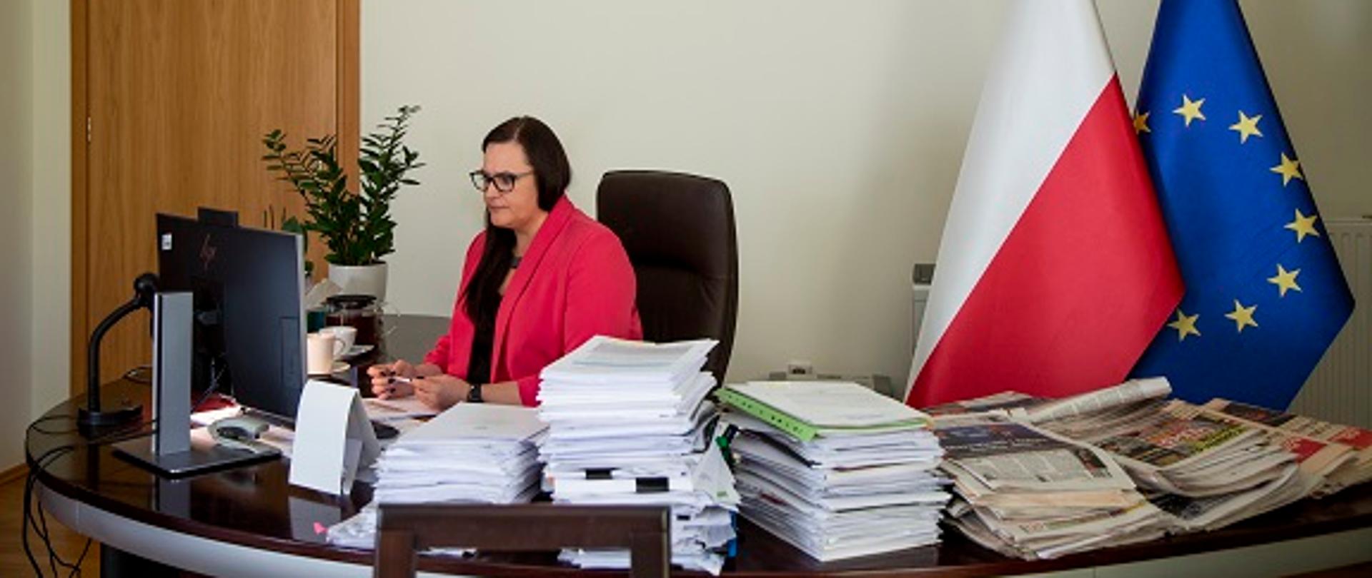 Minister Małgorzata Jarosińska-Jedynka siedzi przy biurku w swoim gabinecie. Za nią flagi Polski i UE. Minister patrzy w komputer, na którym trwa wideokonferencja poświęcona Strategii dla Karpat z udziałem między innymi ratowników górskich z regionu.