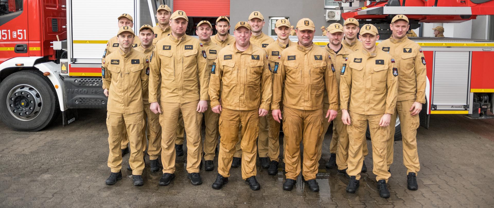 Zdjęcie grupowe strażaków przed drabiną mechaniczną i wozem pożarniczym, cała zmiana ustawiona do wspólnej fotografii przed budynkiem jednostki.