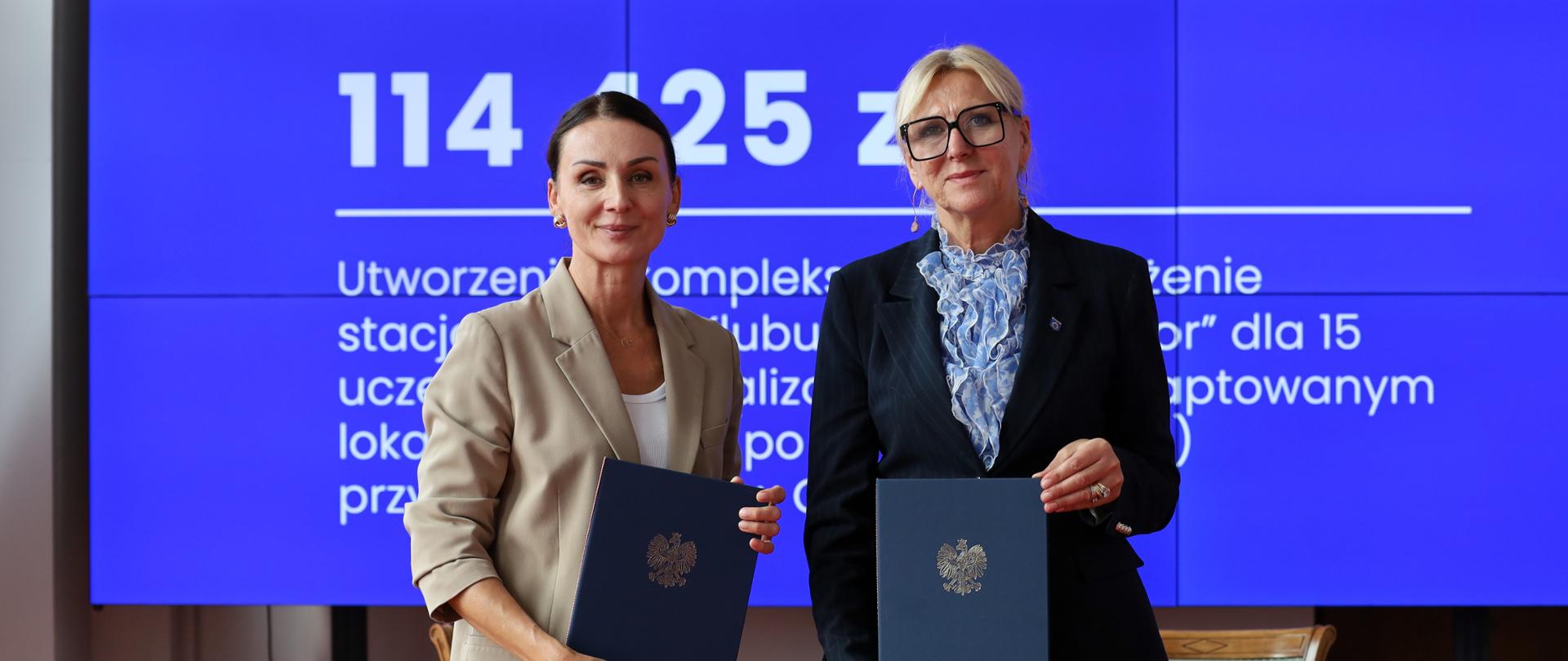 Dwie kobiety stoją obok siebie na tle dużego niebieskiego ekranu prezentacyjnego z napisem „Gmina Gniewino”. Kobieta po lewej stronie ubrana jest w jasną marynarkę, a kobieta po prawej w ciemny garnitur i okulary. Obie trzymają w rękach granatowe teczki z godłem Polski, prezentując podpisane dokumenty.