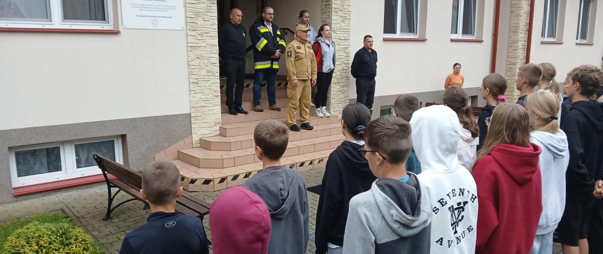 Uczestnicy obozu stoją w dwuszeregu tyłem, na schodach wejściowych do budynku stoją przedstawiciele organizatorów obozu. Na fasadzie budynku flaga Polski