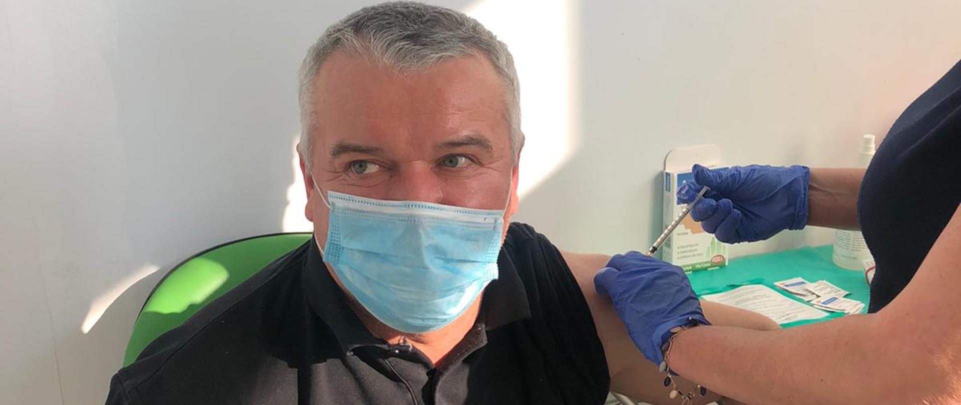 Zdjęcie przedstawia funkcjonariusza PSP (dowódca JRG) oraz personel medyczny podający szczepionkę przeciwko COVID-19 . Zdjęcie wykonane w gabinecie punktu szczepień w Kaliszu w porze dziennej.