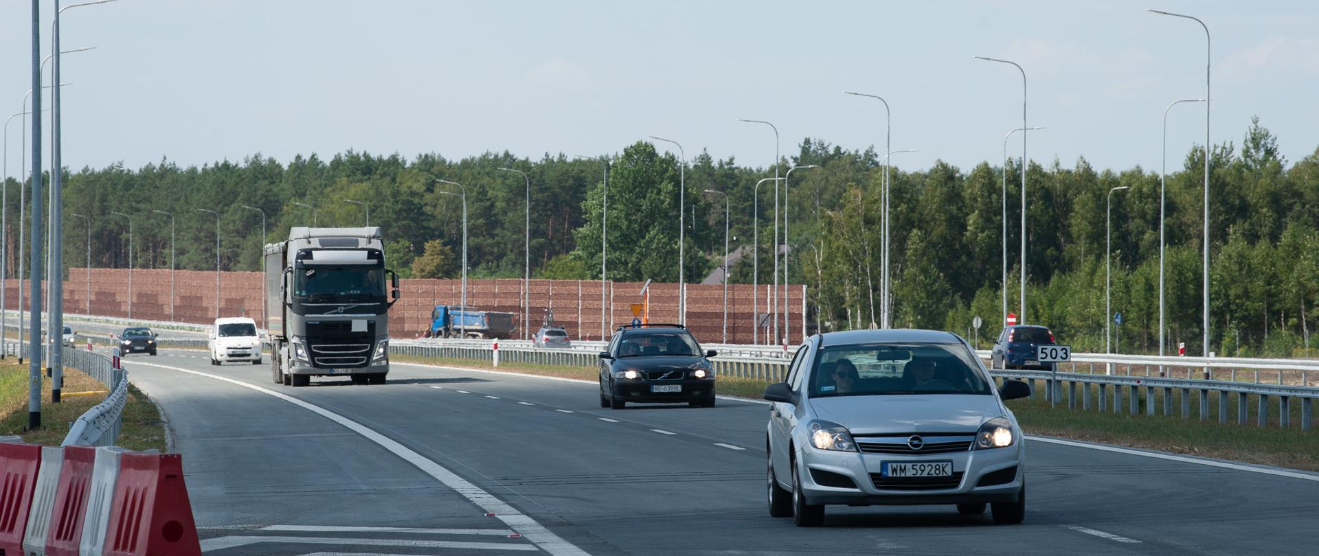 Widok na jezdnię autostrady, po której poruszają się samochody, w tle widoczne ekrany akustyczne i las