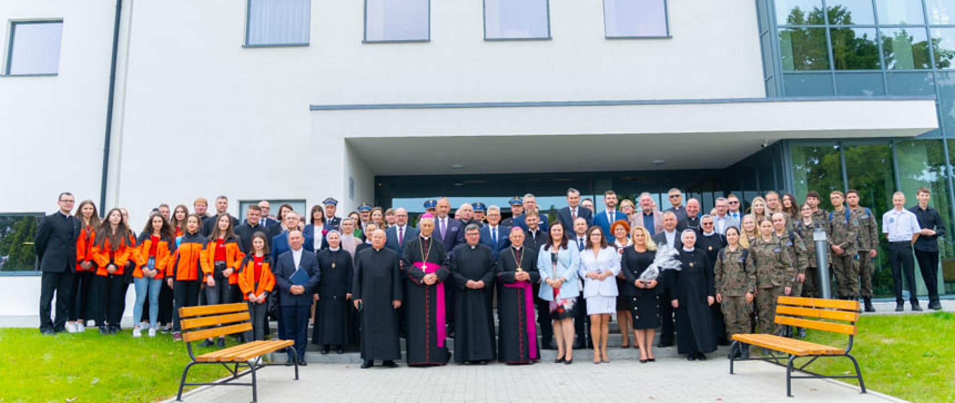 Zdjęcie grupowe uczestników uroczystości otwarcia Centrum Pracy z Młodzieżą - na tle budynku stoją dzieci, młodzież, harcerze, księża, pozostali goście, przedstawiciele administracji, w tym wiceminister Jarosińska-Jedynak.