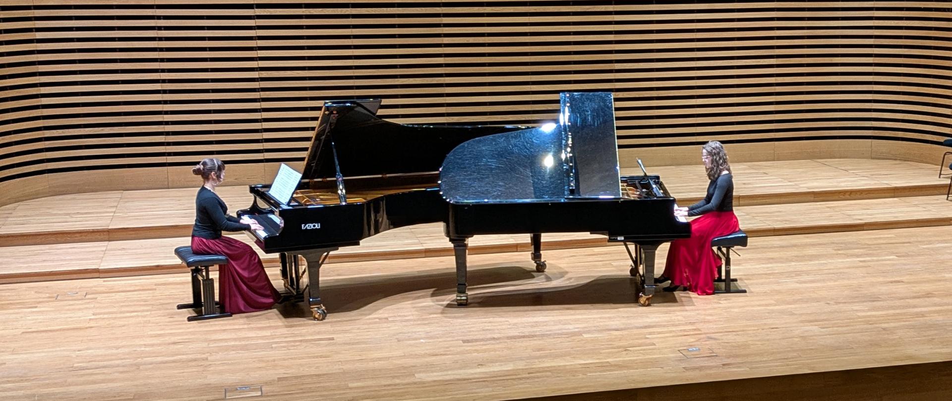 Dwie dziewczyny grają na dwóch fortepianach, ustawionych naprzeciw siebie.