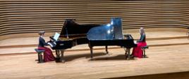 Dwie dziewczyny grają na dwóch fortepianach, ustawionych naprzeciw siebie.