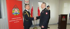 Zdjęcie zrobione w pomieszczeniu strażnicy na tle na tle godła Rzeczpospolitej Polskiej, dwóch Flag Rzeczpospolitej Polskiej oraz baneru Komendy Powiatowej PSP w Lublińcu. Na pierwszym planie Komendant Powiatowy Państwowej Straży Pożarnej w Lublińcu st. bryg. Janusz Bula z uściskiem dłoni z funkcjonariuszem odchodzącym na zaopatrzenie emerytalne. Strażacy w umundurowaniu galowym.