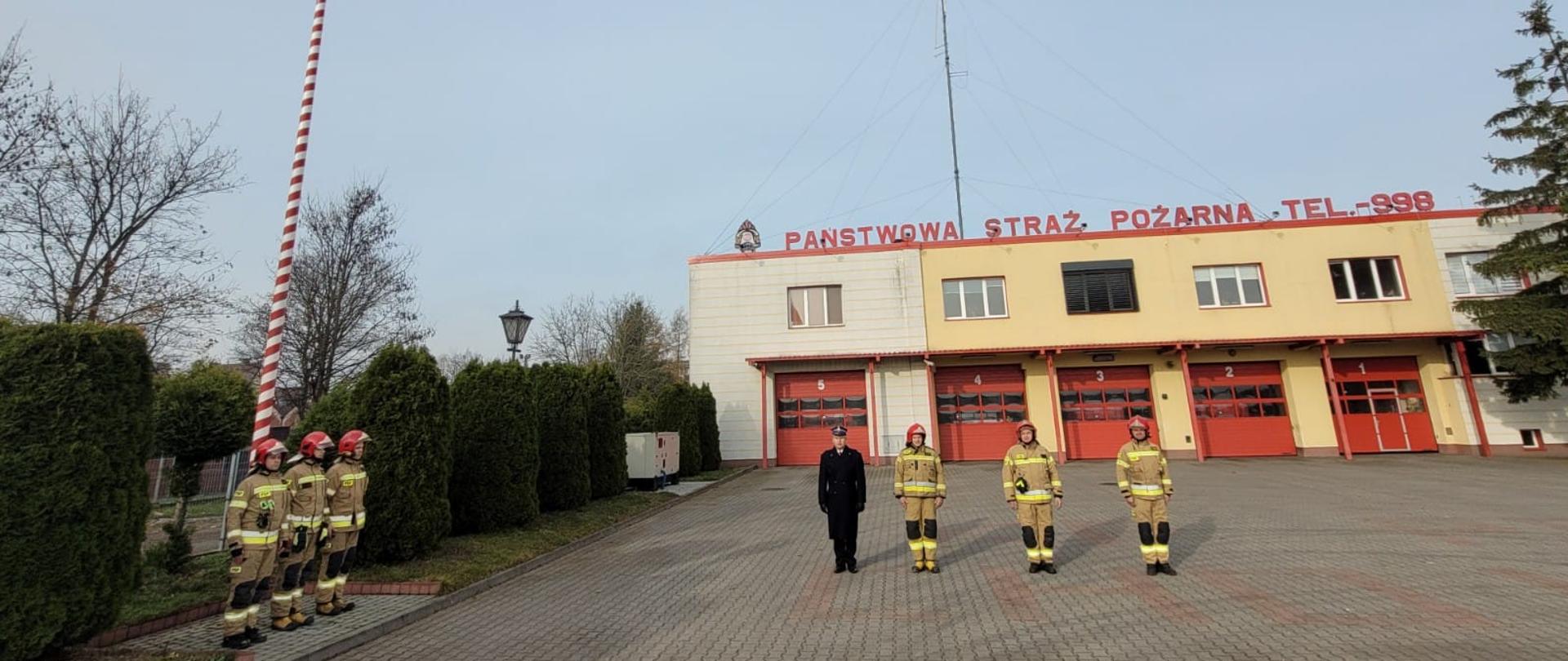 Strażacy podczas uroczystości podniesienia flagi. Stoją na baczność koło masztu flagowego oraz przed budynkiem komendy. Obok tuje rosnące koło ogrodzenia. W tle budynek komendy.