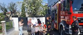 Strażacy Jednostki Ratowniczo – Gaśniczej w Przedszkolu „Bajkowa Kraina” pokazują przez budynkiem dzieciom działanie sprzętu gaśniczego. 