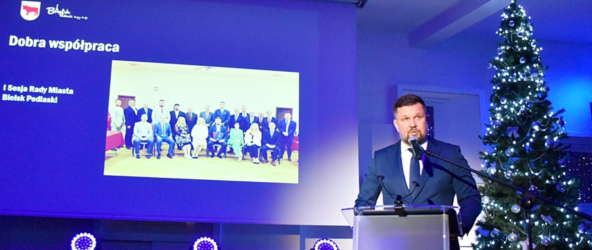 Mównica za którą stoi przemawiający pan. Na niej godło miasta , za udekorowana choinka. W tle duży ekran na którym wyświetlane są zdjęcia.