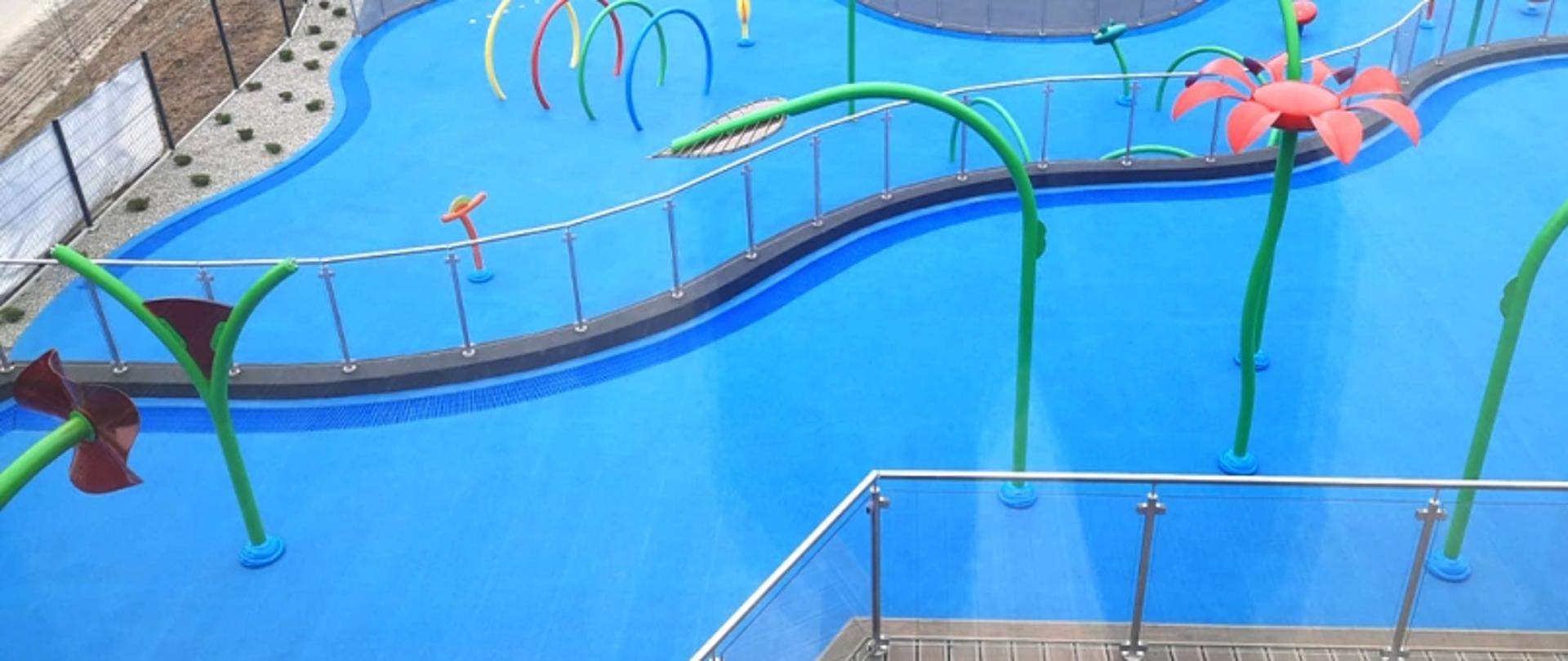 Wodny plac zabaw: kolorowe drabinki, prysznice w kształcie kwiatów i liści.