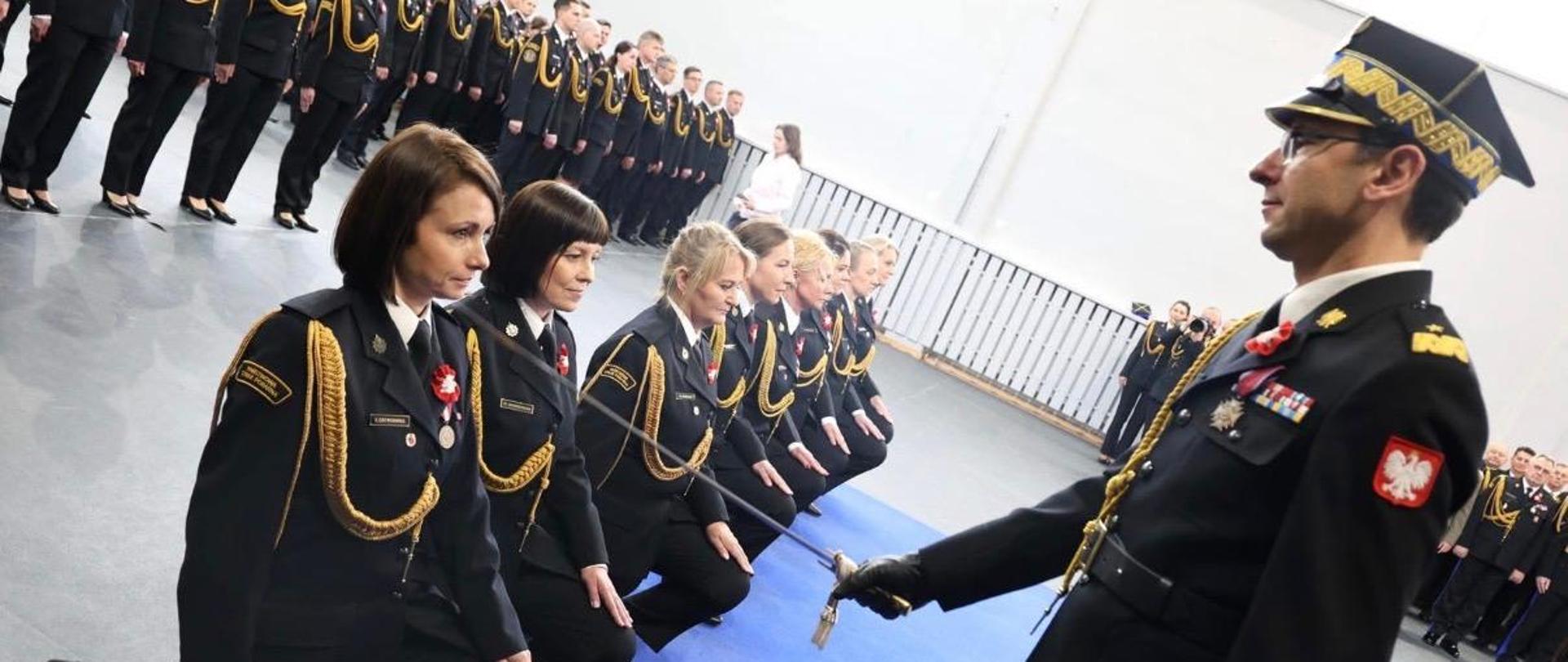 Funkcjonariusz PSP wykonuje pasowanie szablą na klęczących funkcjonariuszach ustawionych w szeregu.