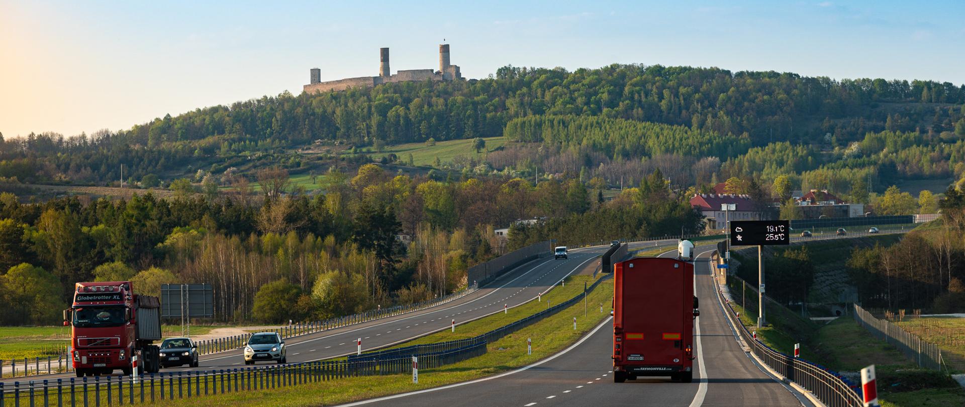 S7 - zamek w Chęcinach. Dwujezdniowa droga ekspresowa S7. W tle na wzgórzu ruiny zamku w Chęcinach. Widoczne trzy baszty otoczone ceglanym murem.