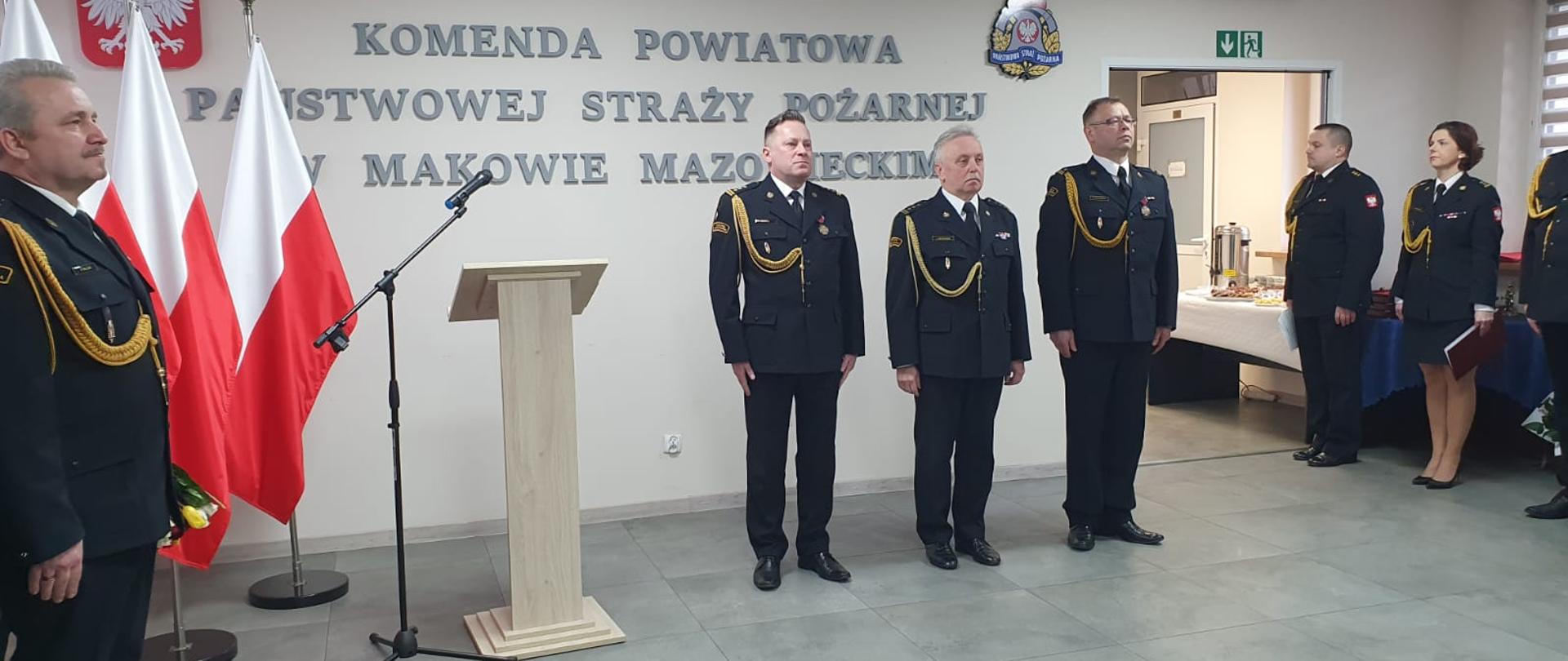 Mazowiecki Komendant PSP oraz Zdający i Przejmujący obowiązki Komendanta Powiatowego stoją na świetlicy podczas uroczystości.
