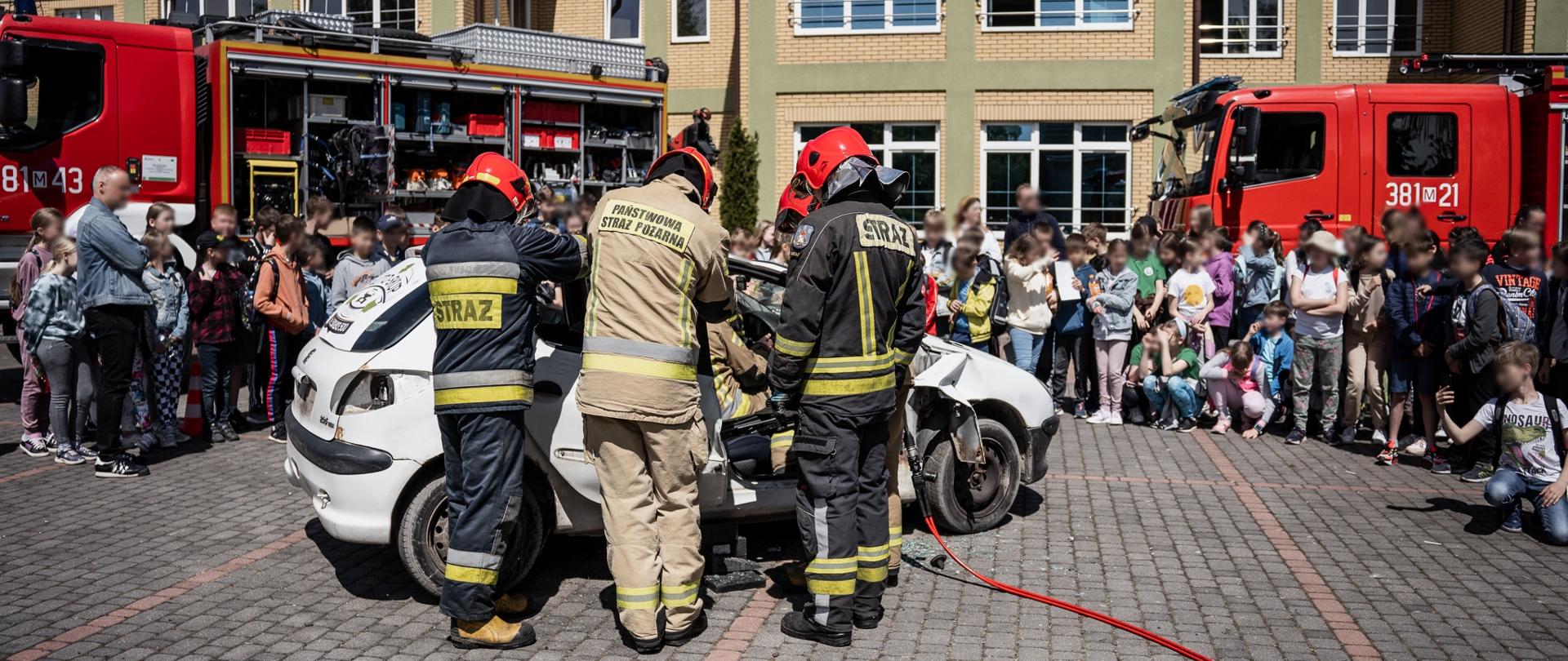 Zdjęcie przedstawia czterech strażaków, ubranych w ubrania specjalne i czerwone hełmy, którzy sprzętem hydraulicznym wykonują dostęp do osoby poszkodowanej, znajdującej się we wraku samochodu. W tle zdjęcia grupa dzieci obserwujących pokaz, na tle dwóch samochodów pożarniczych. 
