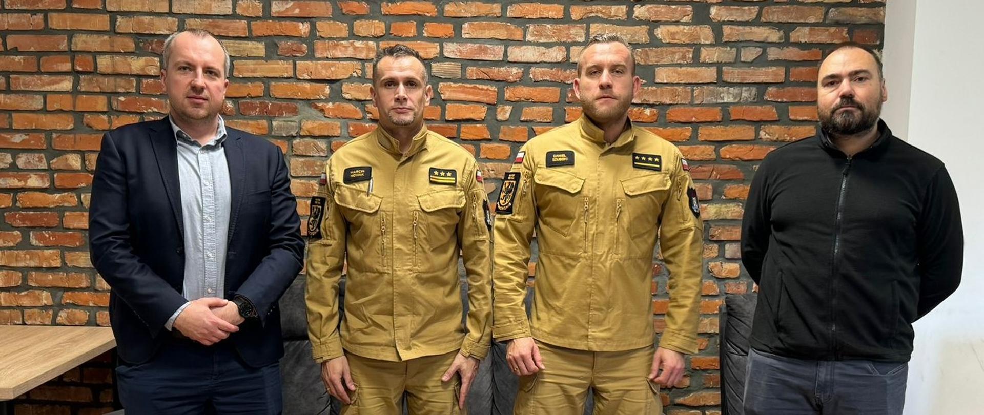 Przy stole konferencyjnym kadra dowódcza stoi z przedstawicielami firmy. Drewniane meble oraz ściana z starych czerwonych cegieł podkreśla niesamowity klimat wydarzenia jakim jest przekazanie komputera dla PSP.