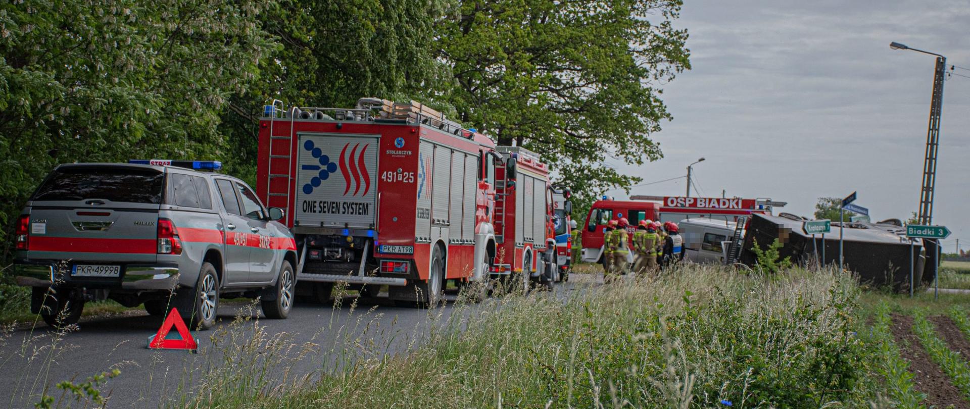 Wypadek dwóch samochodów. Zdjęcie przedstawia samochody służb biorących udział w zdarzeniu oraz rozbite samochody. Strażacy stoją przy samochodach.