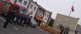 Strażacy stoją w pozycji zasadniczej, dowódcy i poczet flagowy oddają honor wciąganej na maszt Flagi Państwowej RP. Przed budynkiem komendy stoją wozy strażackie.