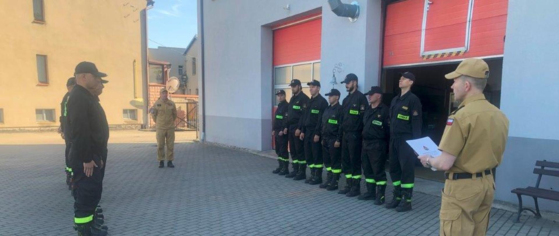 Zdjęcie wykonano w ciągu dnia na placu Komendy Powiatowej Państwowej Straży Pożarnej w Sępólnie Krajeńskim, strażacy w ubraniach koszarowych stoją w szeregu naprzeciw siebie, po środku z prawej strony stoi starszy brygadier Maciej Suchomski w mundurze służbowym, a z lewej dowódca Jednostki Ratowniczo-gaśniczej młodszy brygadier Robert Liss.