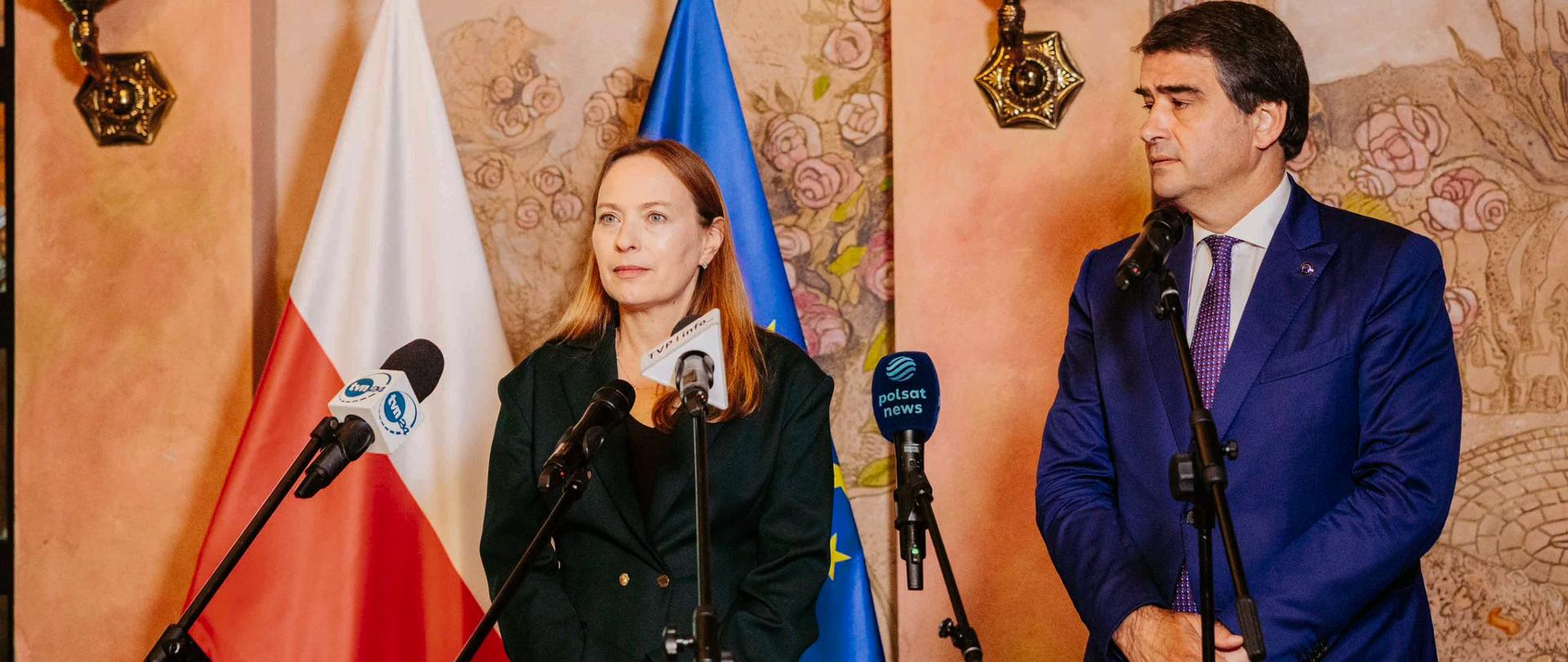 Minister Katarzyna Pełczyńska-Nałęcz oraz wiceprzewodniczący KE Raffaele Fitto stoją przed mikrofonami na tle flag.