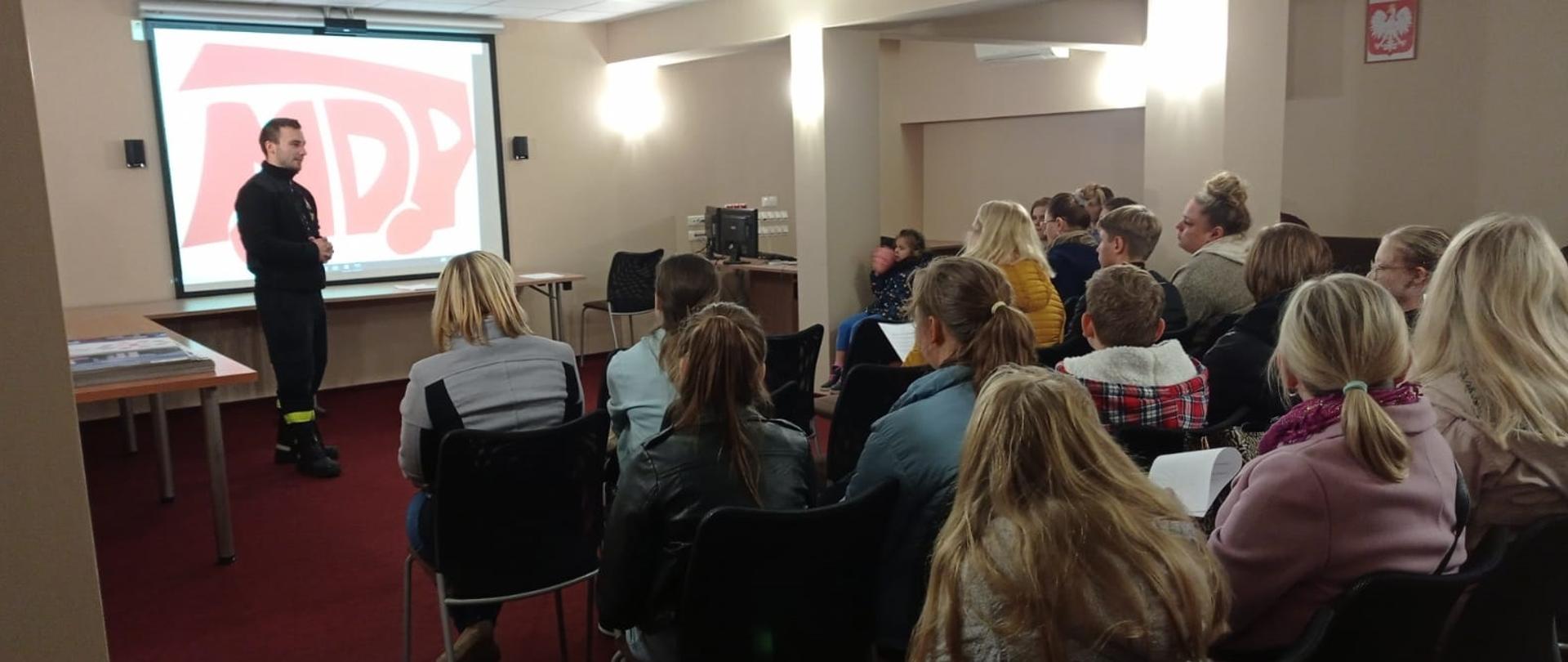 W pomieszczeniu na krzesłach tyłem siedzi kilkadziesiąt osób. Przed nimi obok ekranu stoi strażak w czarnym mundurze.