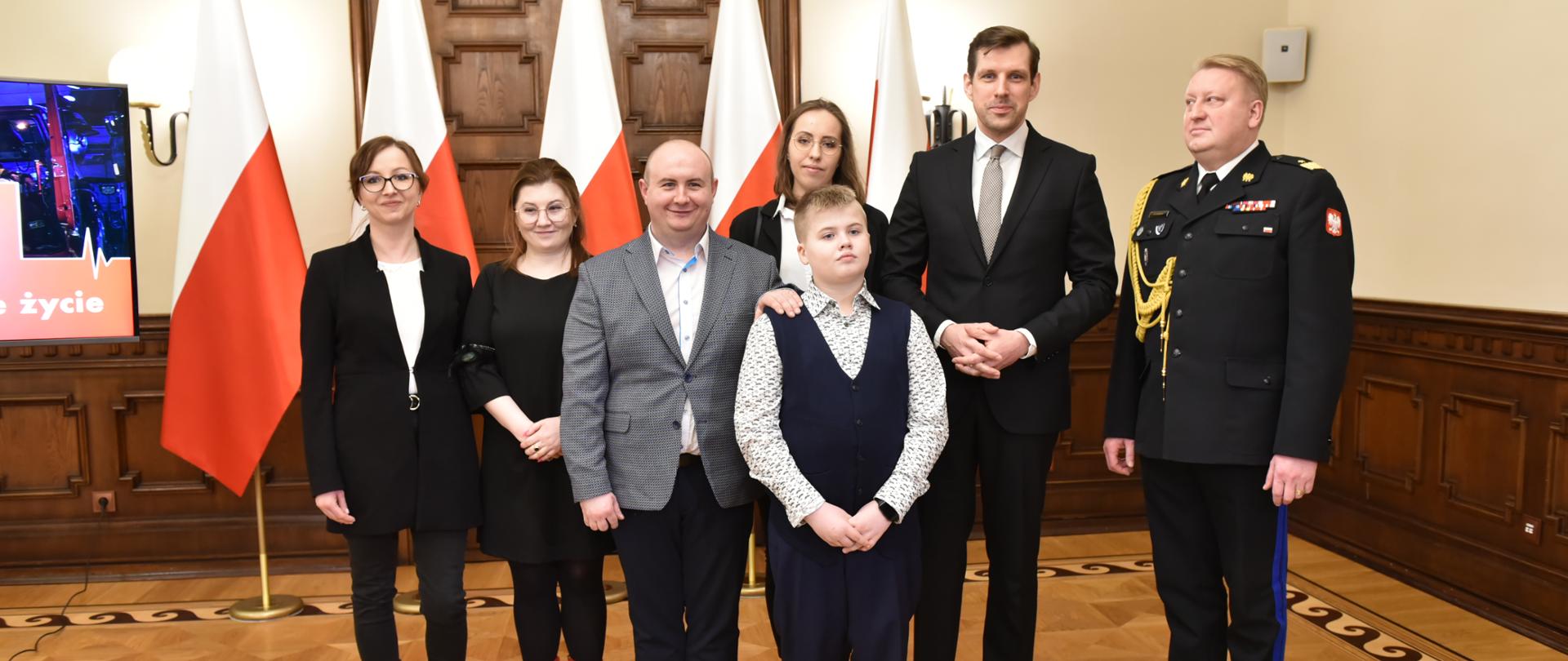 komendant wojewódzki, wojewoda łódzki chłopiec z rodzicami i dwie kobiety pozują do zdjęcia w czasie konferencji prasowej