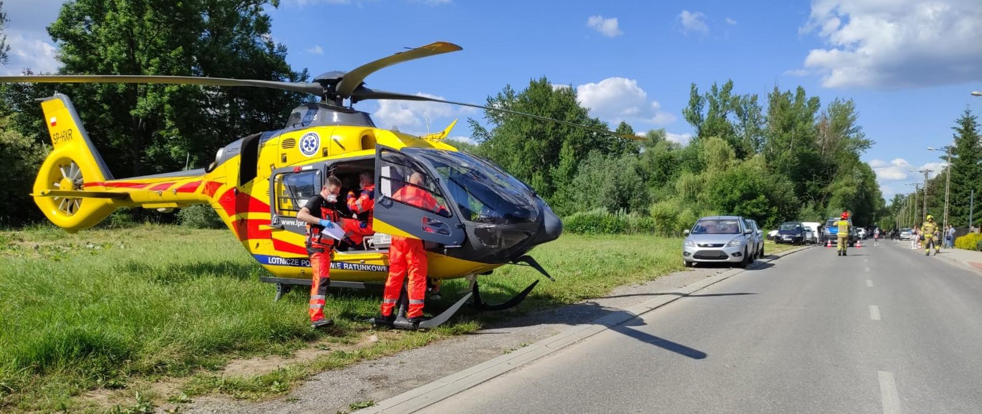 Na zdjęciu widzimy drogę powiatową, która została zamknięta dla ruchu. Na głównym planie śmigłowiec LPR i ratownicy udzielający pomocy osobie poszkodowanej. Samochody osobowe w związku z zatrzymaniem ruchu stoją zaparkowane na poboczu i na jezdni. 
