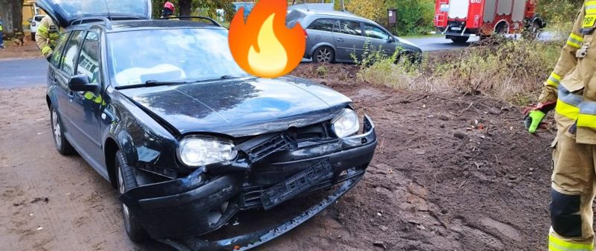 Widok na rozbite auto. Auto koloru czarnego. Ma zniszczony przód,uszkodzona jest maska, grill i lampy.