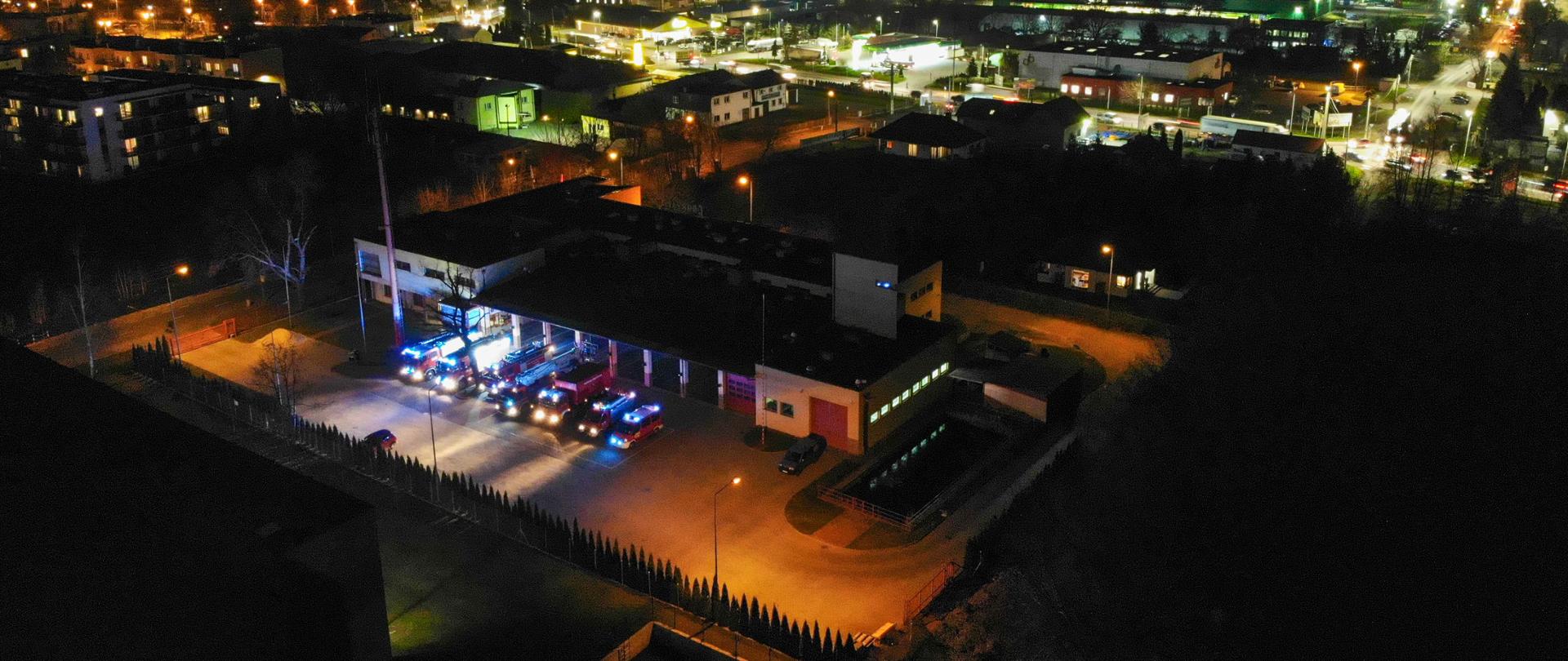 Widok na KP PPS w Grodzisku Maz. z lotu ptaka w nocy. Przed garażami stoją samochody pożarnicze z włączonymi sygnałami błyskowymi. W tle miasto rozświetlone światłami.