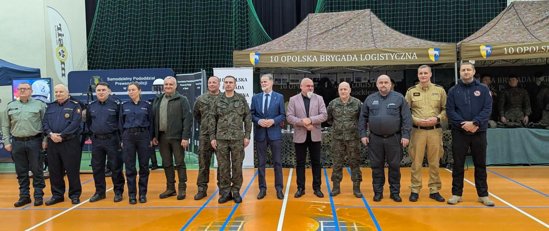 Wspólne zdjęcie przedstawicieli służb mundurowych i samorządu podczas wydarzenia „Spotkanie z mundurem”; w tle stoiska wojskowe i policyjne.