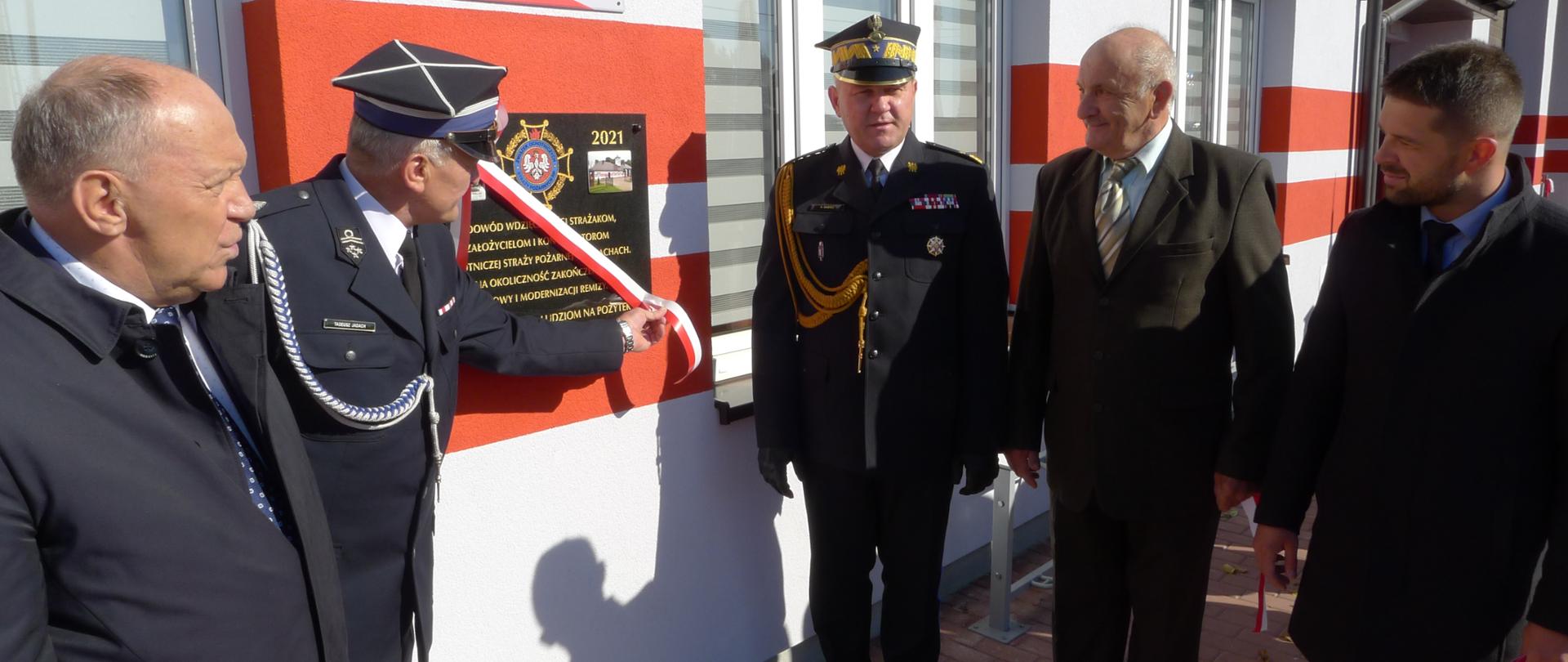 Przed tablicą pamiątkową zamocowaną w ścianie budynku stoi pięciu mężczyzn. Jeden z nich trzyma za biało-czerwona wstęgę zamocowaną do tablicy