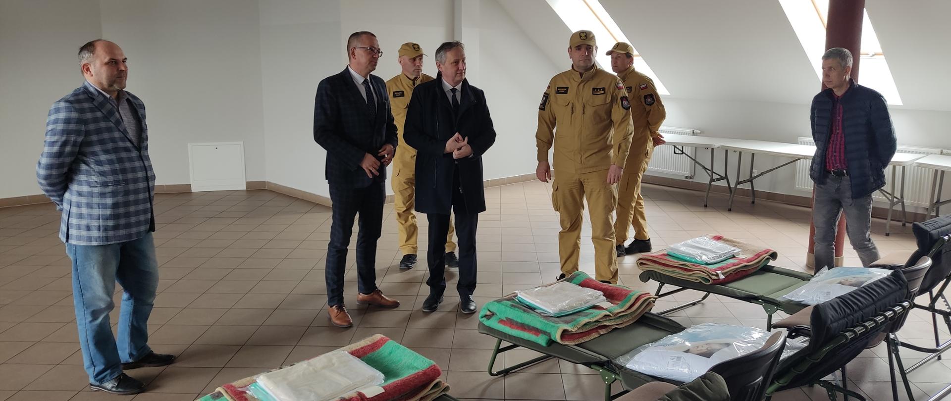 Na zdjęciu widzimy siedmiu mężczyzn rozmawiających wewnątrz sali przystosowanej dla uchodźców z Ukrainy. Widzimy również łóżka polowe oraz krzesła przygotowane dla potrzebujących.