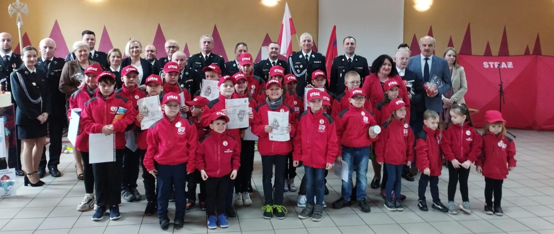 Grupowe zdjęcie, z przodu stoją dzieci wszystkie ubrane w czerwone bluzy , za nimi stoją strażacy i goście cywilni