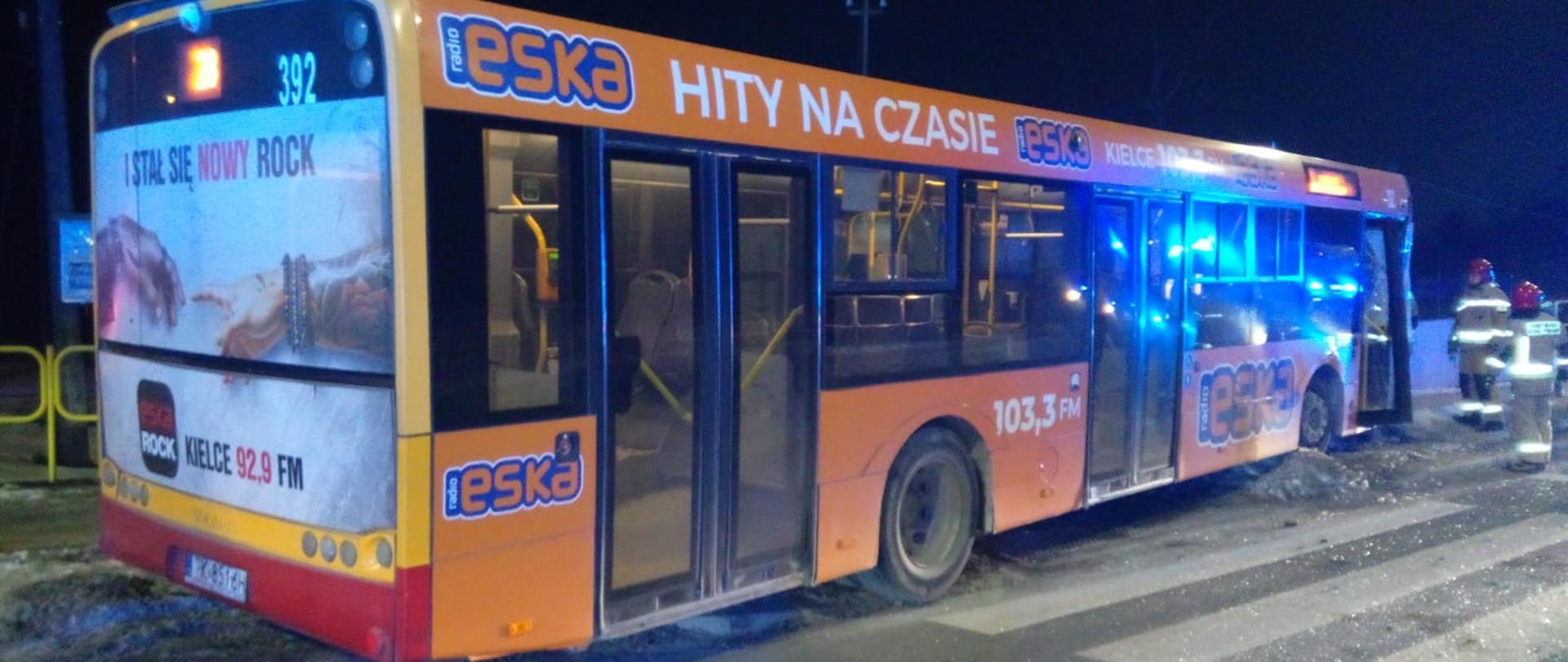 Zdjęcie przedstawia autobus miejski, który uległ zderzeniu. Przy pojeździe stoją strażacy w mundurach.