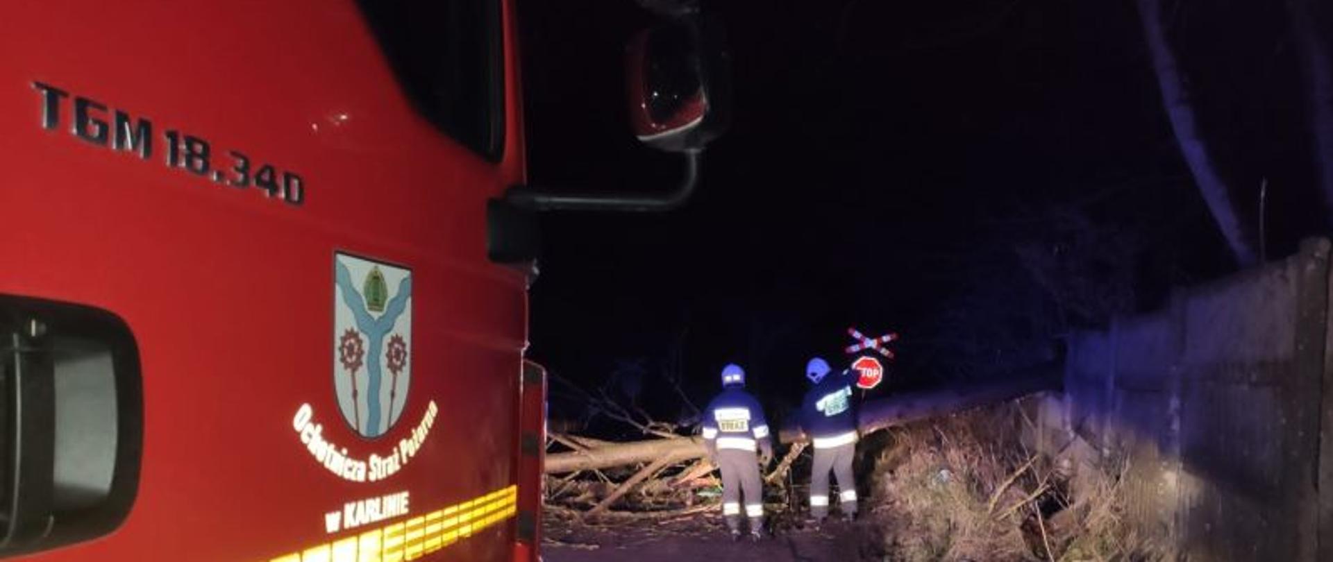Zdjęcie wykonane w nocy. Po lewej stronie zdjęcia widoczny czerwony bok pojazdu gaśniczego. Na drzwiach widnieje logo OSP Karlino. W oddali, oświetleni żółtym światłem pojazdu strażacy. Strażacy w białych hełmach pracują przy leżącym na jezdni drzewie