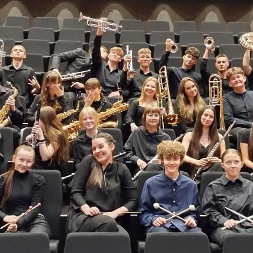 Orkiestra dęta wraz z Panią Dyrygent siedzą w kilku rzędach na widowni sali koncertowej, wszyscy trzymają w rękach swoje instrumenty.