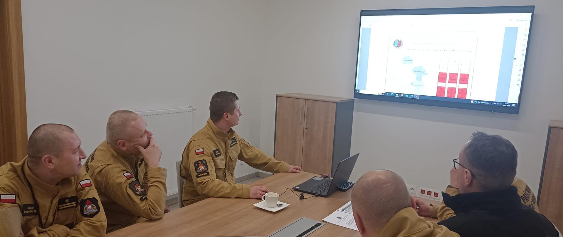 Zdjęcie przedstawia sześciu funkcjonariuszy PSP siedzacych przy stole. na ścianie znajduje się ekran