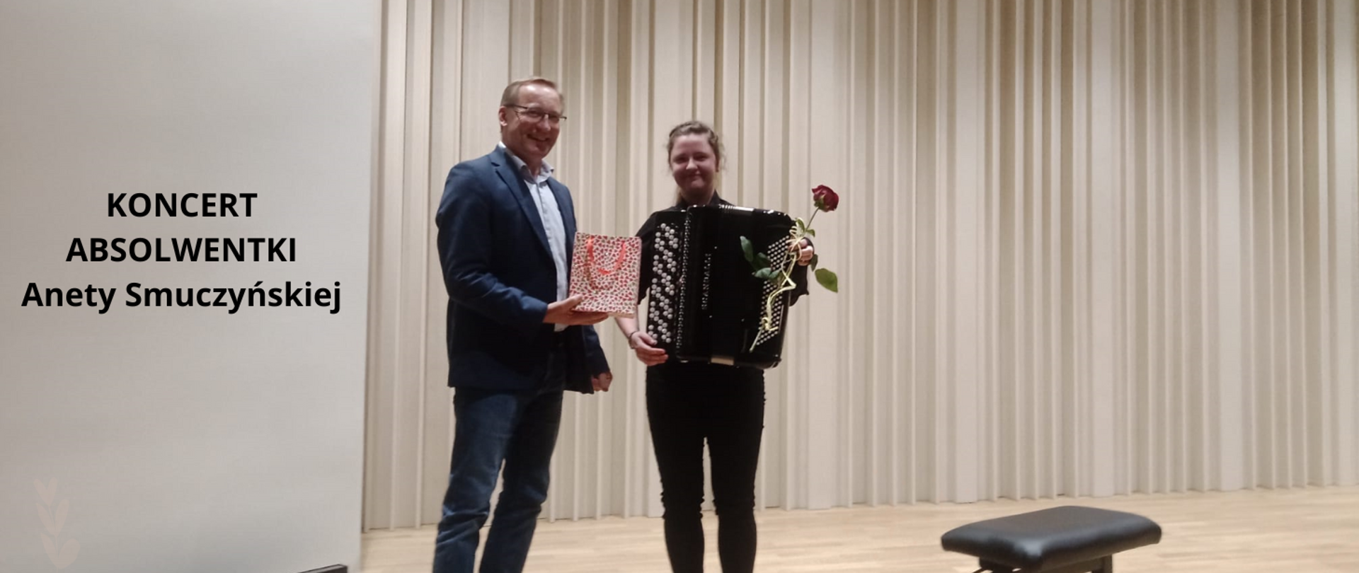 Na zdjęciu w centralnej części obrazka dwie osoby. Po lewej mężczyzna w granatowej marynarce i białej koszuli, trzyma w ręku torebkę prezentową. Po prawej stronie na czarno ubrana dziewczyna z akordeonem, która trzyma czerwoną różę. Po lewej stronie obrazka czarny napis: "koncert absolwentki Anety Smuczyńskiej". W tle akustyczne panele w odcieniach brązu.