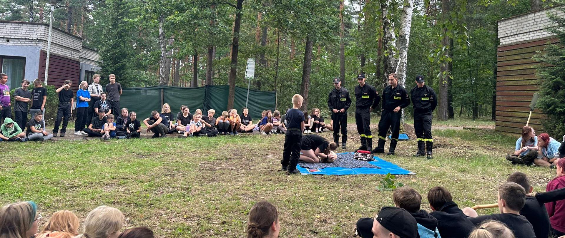  W otoczeniu lasu dzieci i młodzież siedzą na ziemi na planie kwadratu. Przed nimi czterech funkcjonariuszy Państwowej Straży Pożarnej oraz manekin do nauki udzielania pierwszej pomocy. Obok manekina chłopiec i dziewczyna ćwiczący udzielanie pierwszej pomocy.