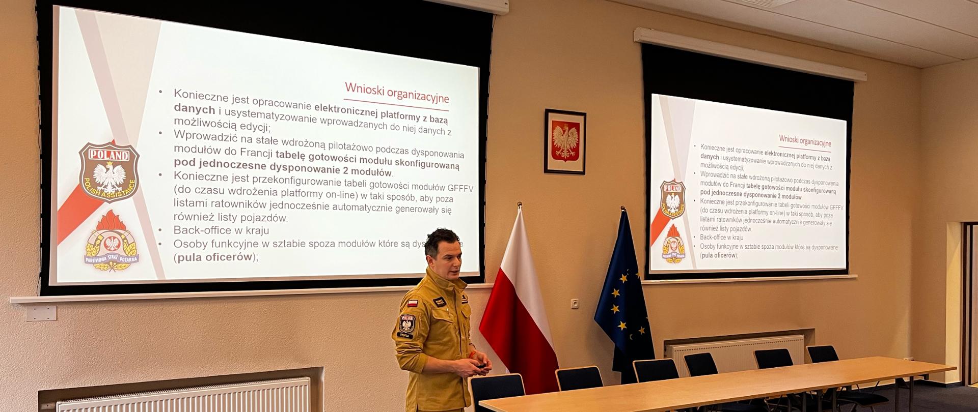 Oficer PSP w mundurze służbowym koloru piaskowego prowadzi wykład, za nim na ścianie wyświetlony jest slajd z treścią przekazu oraz logo PSP i modułu GFFFV