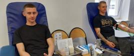 Na zajęciu widoczni dwaj mężczyźni obaj w czarnych koszulkach i szarych spodniach siedzący na fotelach na sali oddający krew podczas akcji zbiórki krwi zorganizowanej przez Klub Honorowych Dawców Krwi Polskiego Czerwonego Krzyża przy Komendzie Powiatowej Państwowej Straży Pożarnej w Gorlicach. 