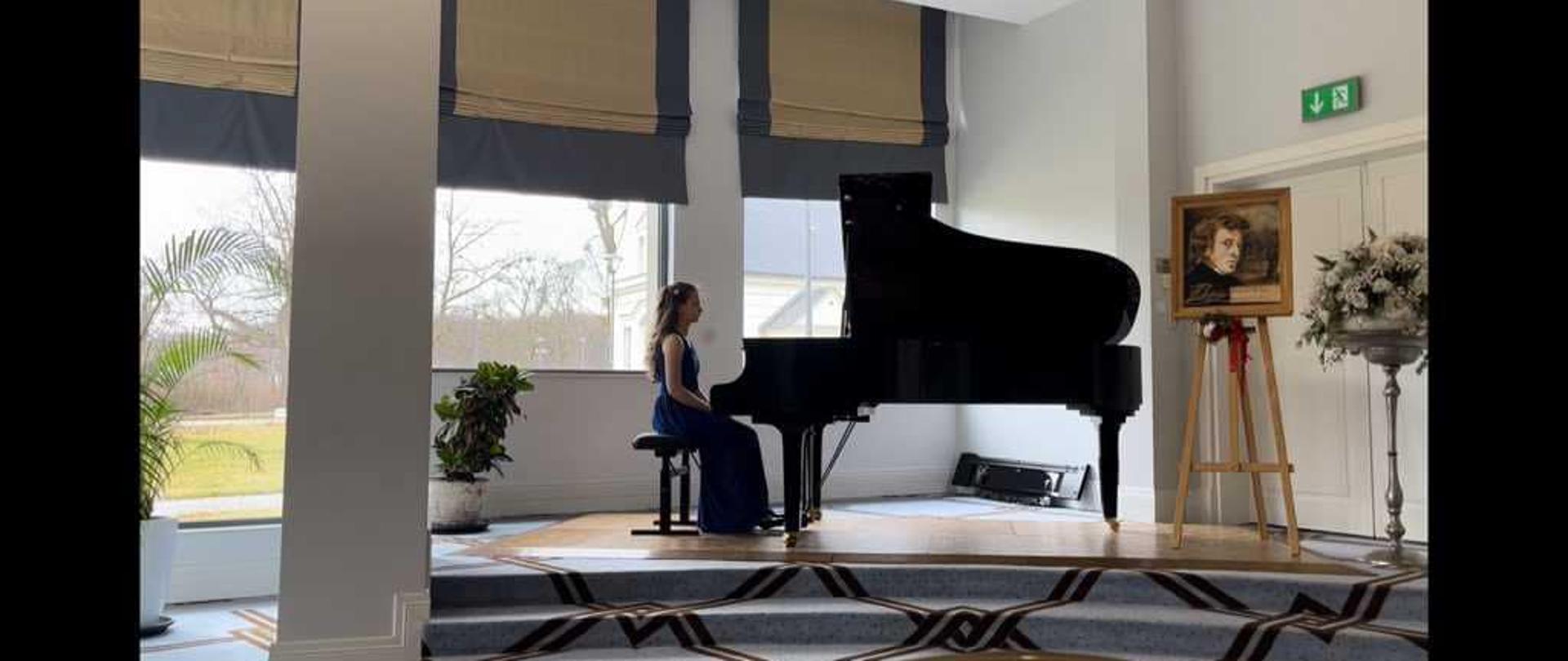 Pianistka siedzi przy fortepianie, w tle okna na ogórd, po prawej stronie portret Chopina na sztaludze i kwiaty, dywan w niebiesko-brązową kratę. 