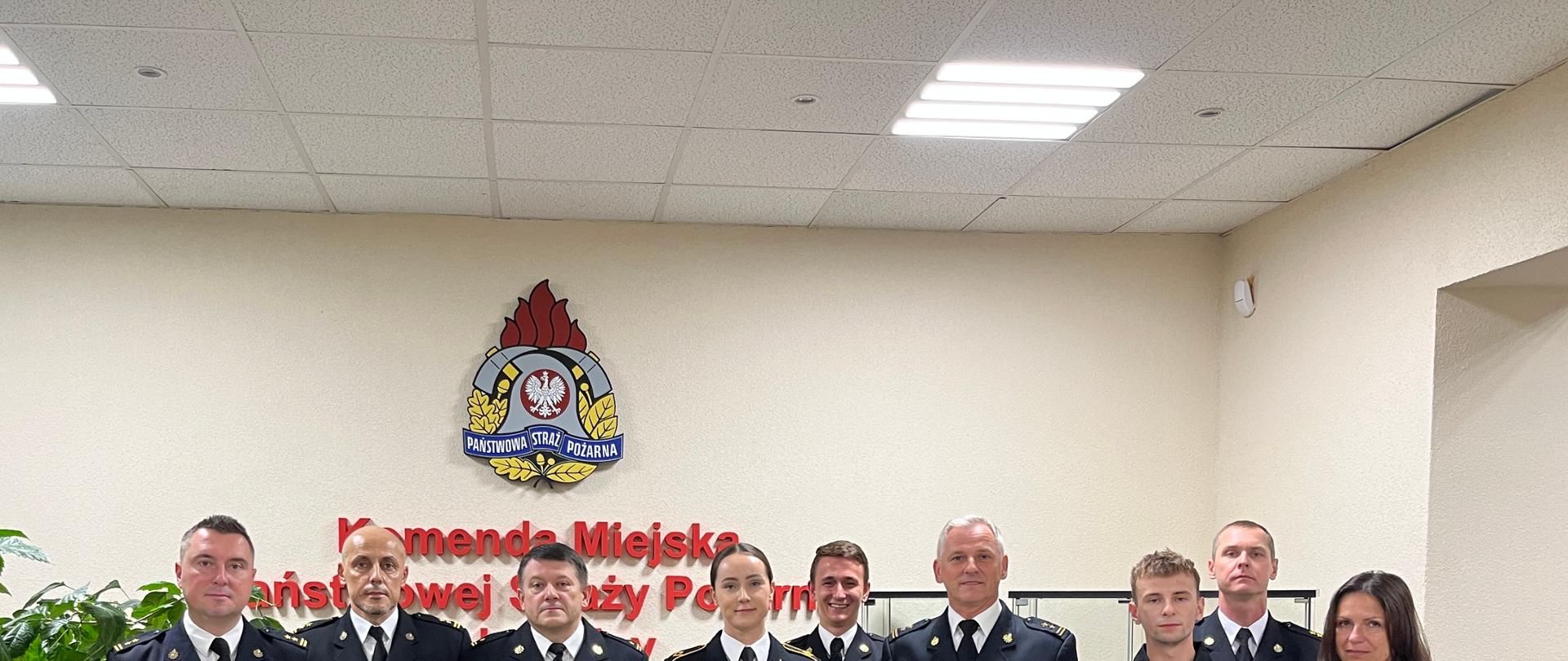 Wszyscy uczestnicy stoją w jednym rzędzie na wprost aparatu. W grupie dziewięciu osób znajdują się zarówno przyjęci wcześniej młodzi funkcjonariusze, jak i wyższa kadra oficerska w galowych mundurach wyjściowych, ozdobionych baretkami odznaczeń. W tle, centralnie nad nimi, widoczne jest duże logo Państwowej Straży Pożarnej oraz czerwony napis Komenda Miejska Państwowej Straży Pożarnej w Legnicy.