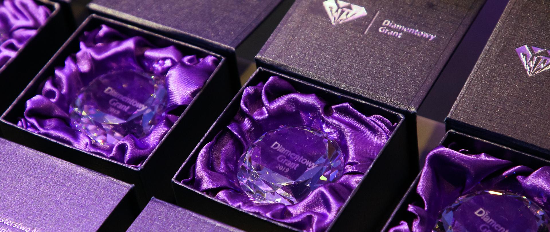 Fioletowe statuetki przedstawiające diamentowe granty. Diamenty leżą w pudełeczkach wyściełanych materiałem.