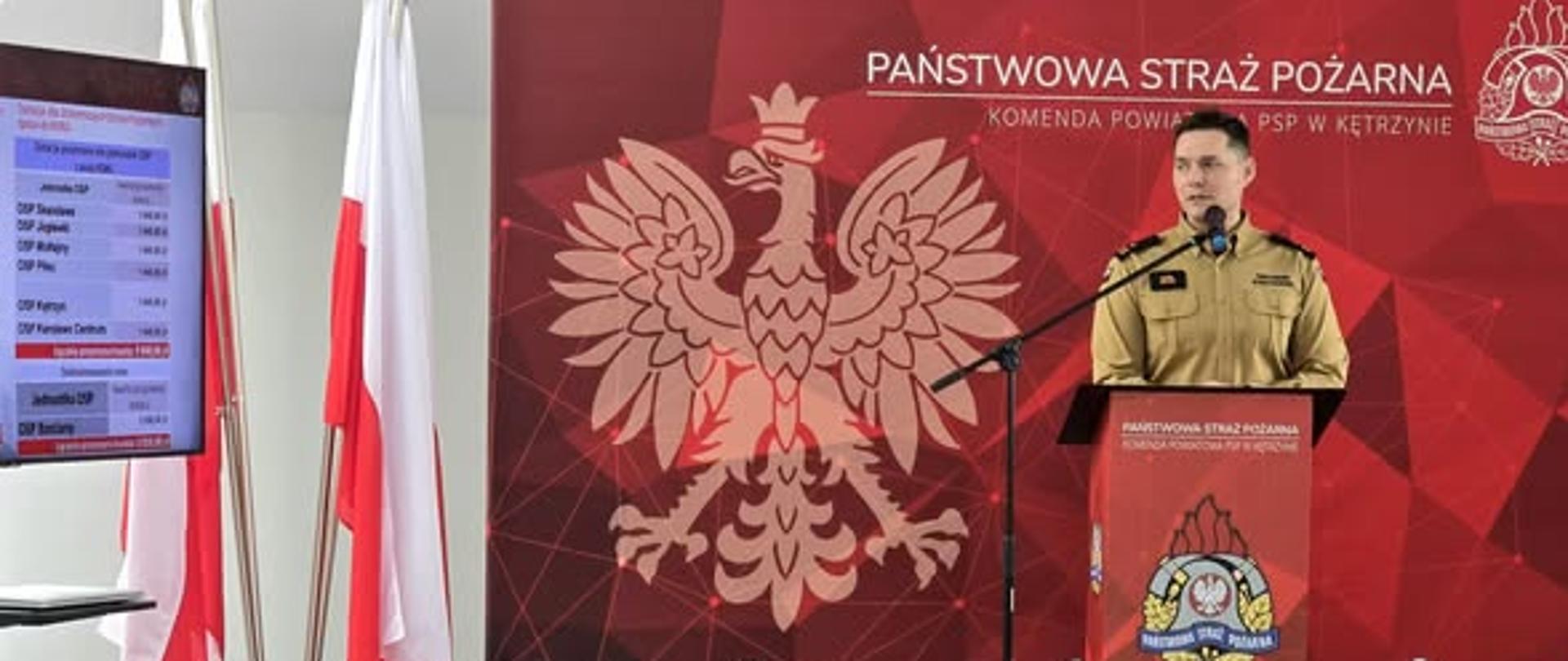 Na zdjęciu widać funkcjonariusza w żółtym ubraniu służbowym, stojący przy mównicy koloru czerwonego z logo Państwowej Straży Pożarnej. W tle flaga Polski oraz telewizor. Z tyłu ścianka koloru czerwonego z logo Państwowej Straży Pożarnej.