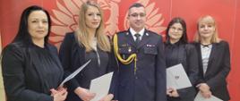 Komendant Miejski wraz z pracownicami Komendy stoją na tle ścianki firmowej. Komendant w środku. Panie ubrane w czarne garsonki trzymają w rękach teczki firmowe.
Nad ich głowami na ściance napis Państwowa Straż Pożarna. 