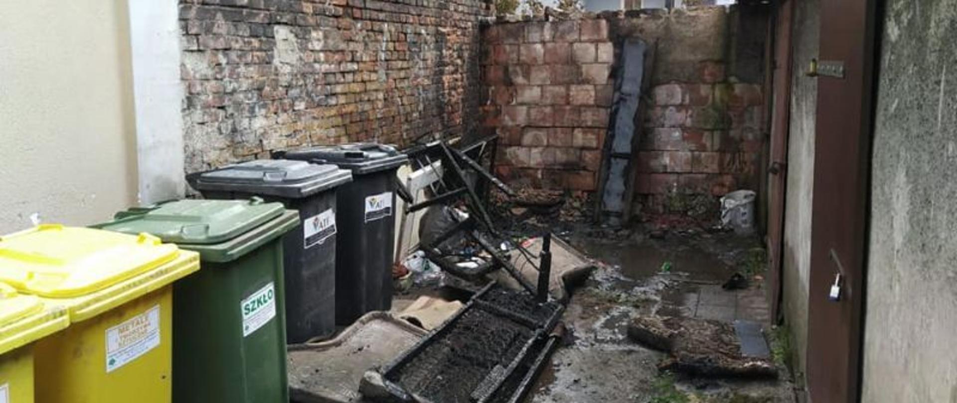 Widok na małe podwórko. Pod ścianą stoją śmietniki do segregacji odpadów. Przy śmietnikach pozostałości po spalonej wersalce. Kawałki stelarzu i sprężyn.